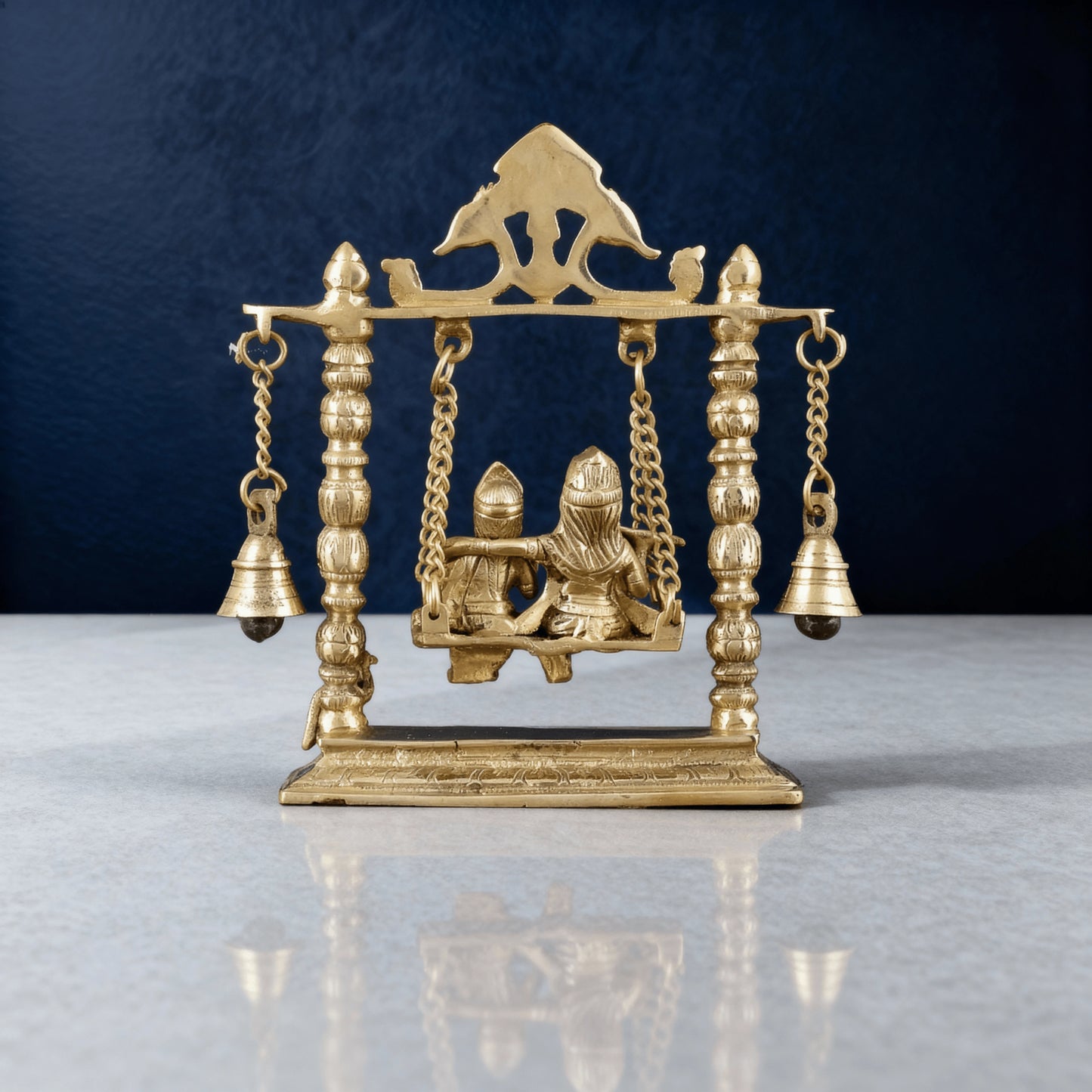 Pure brass Radha Krishna swing idol with intricate bells and pillars on marble surface, handcrafted traditional Indian décor piece.