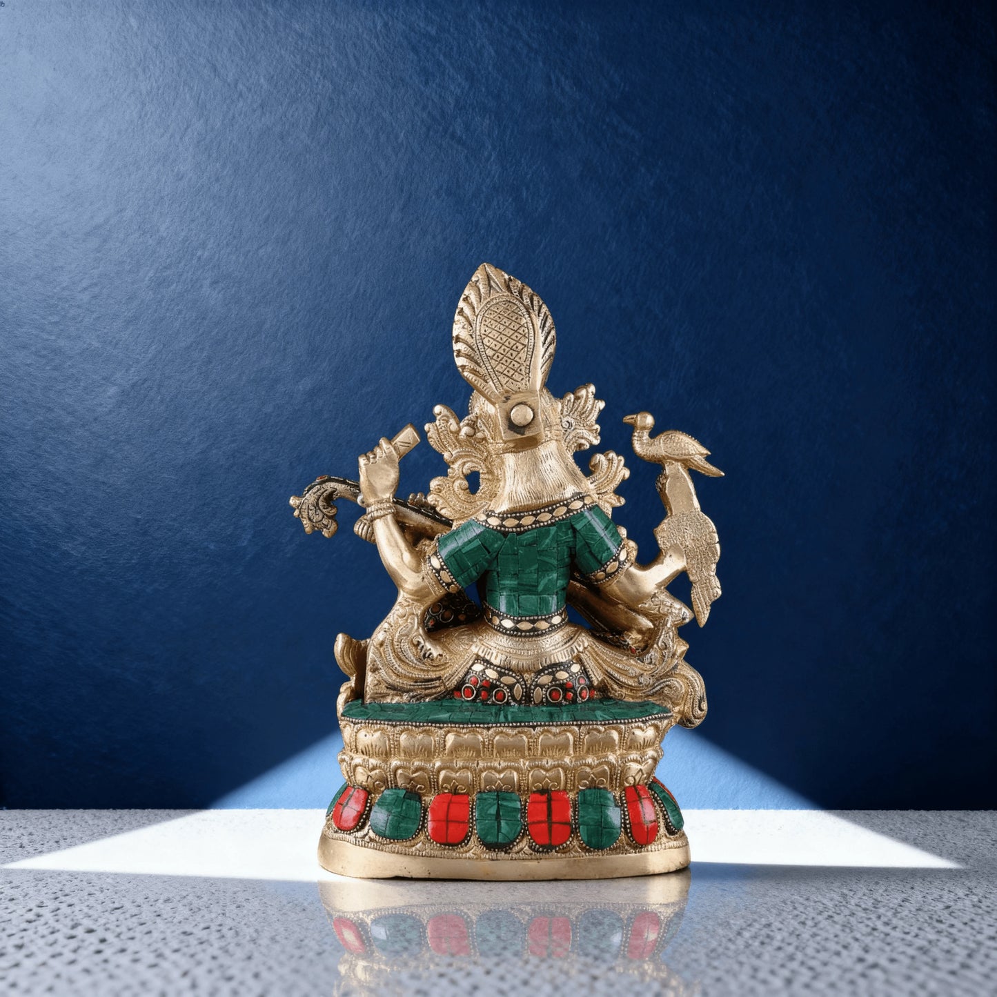 rear view of brass Saraswati statue displaying green and red stonework on the back and base, photographed in studio lighting