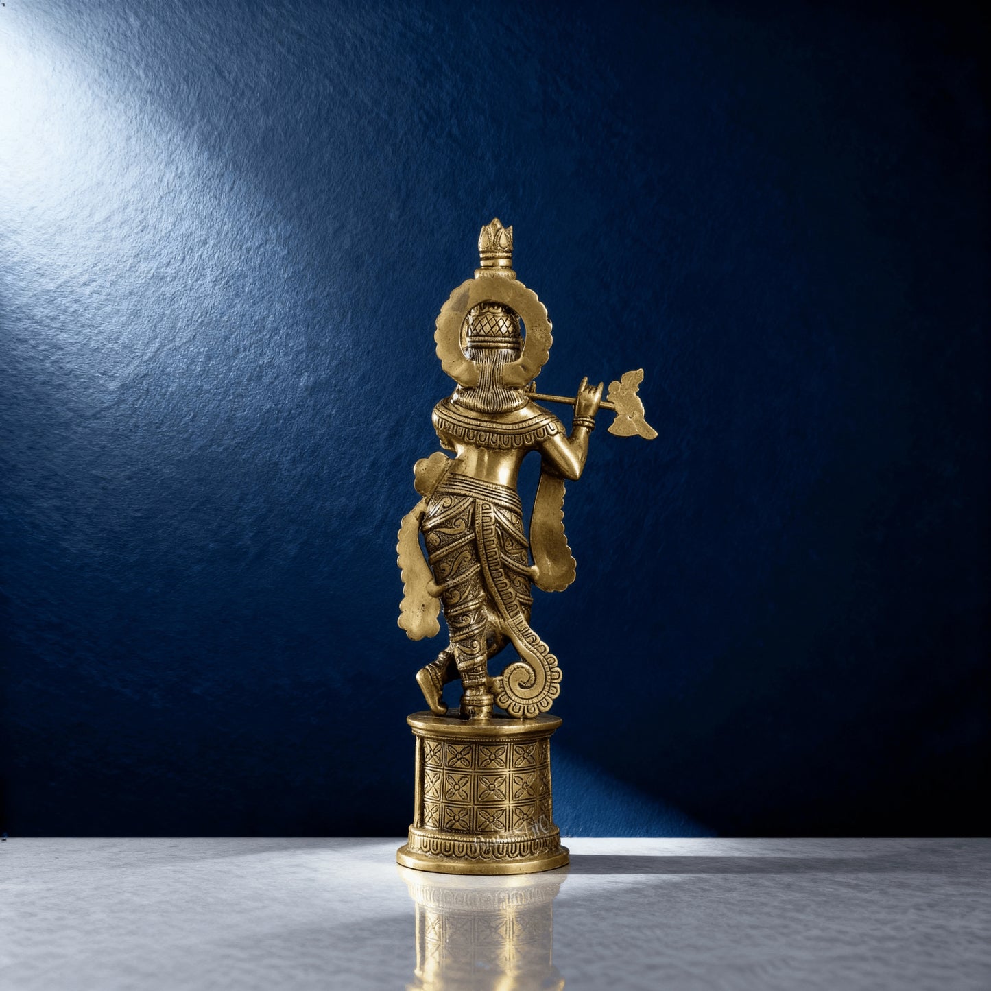 Rear view of polished brass Krishna sculpture showing intricate carvings and flowing drape, placed on marble surface with blue studio background.