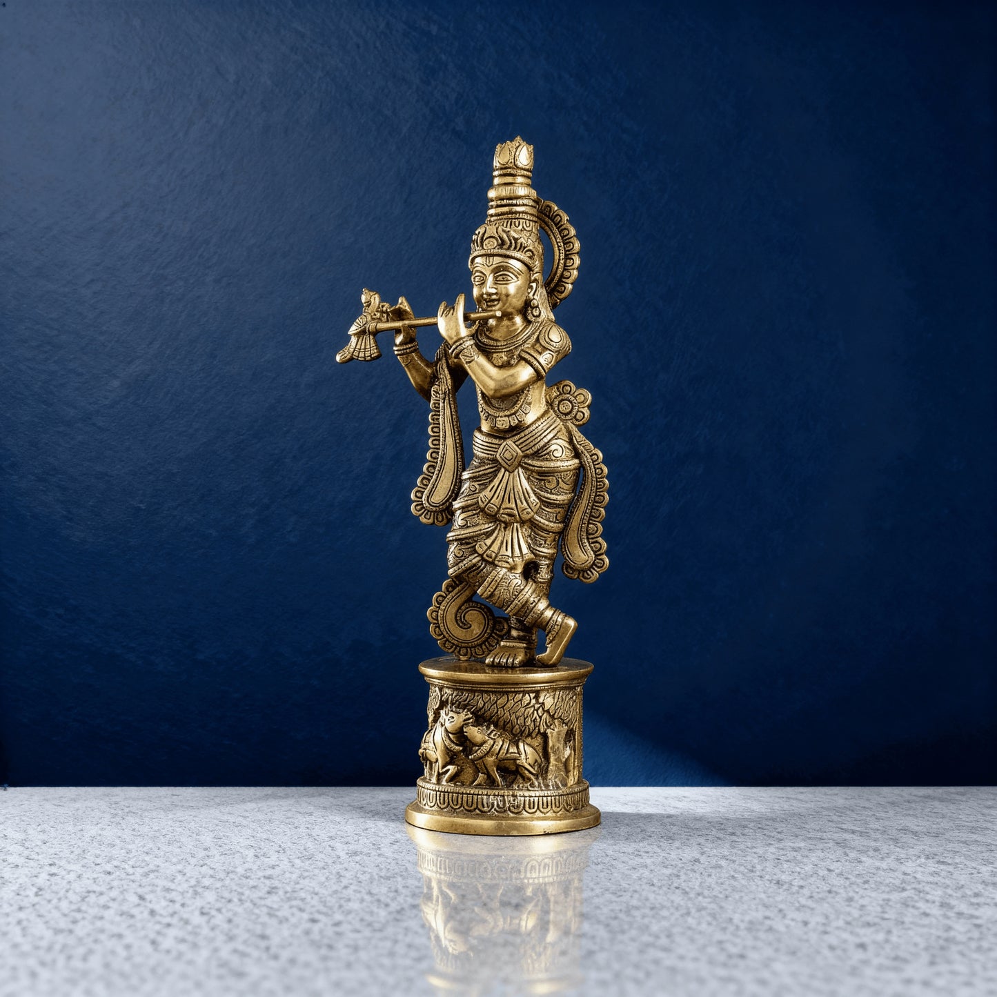 Handcrafted brass Krishna idol with flute and ornate detailing, standing on engraved base, displayed on textured marble with deep blue backdrop.