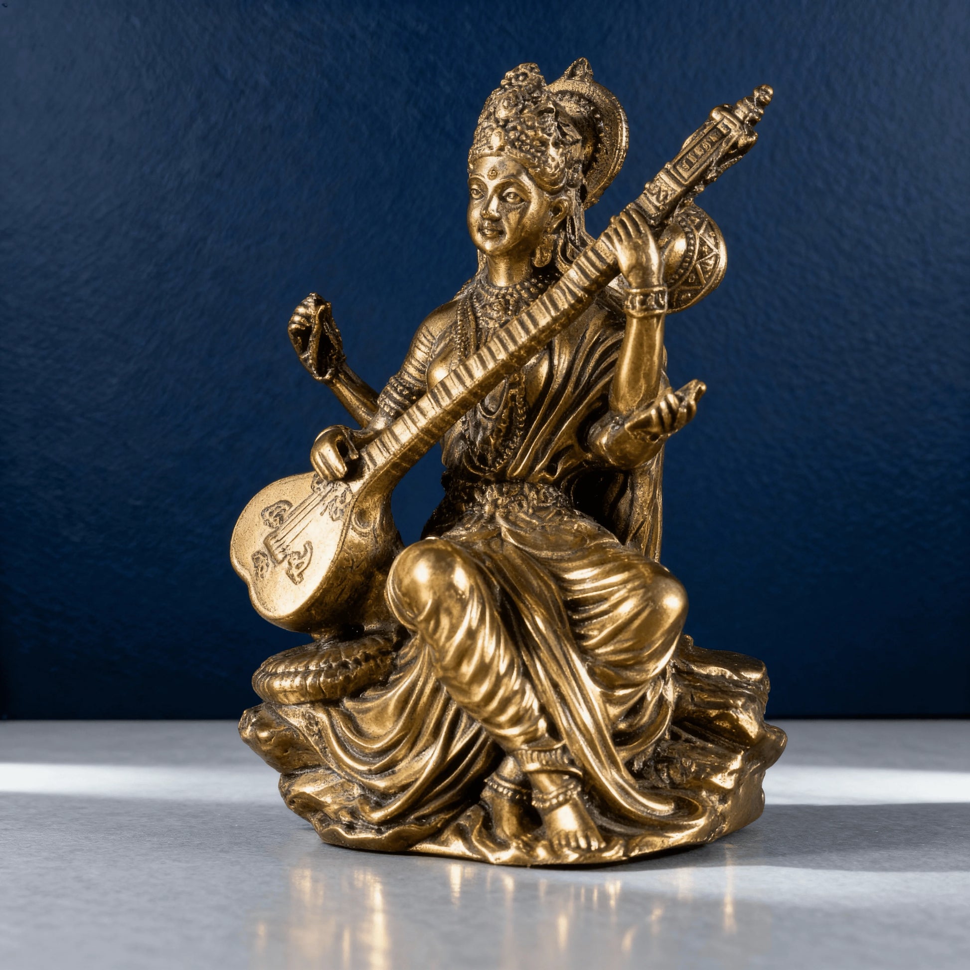 brass Saraswati idol playing the veena, captured in soft studio lighting with a dark blue background and detailed carving visible
