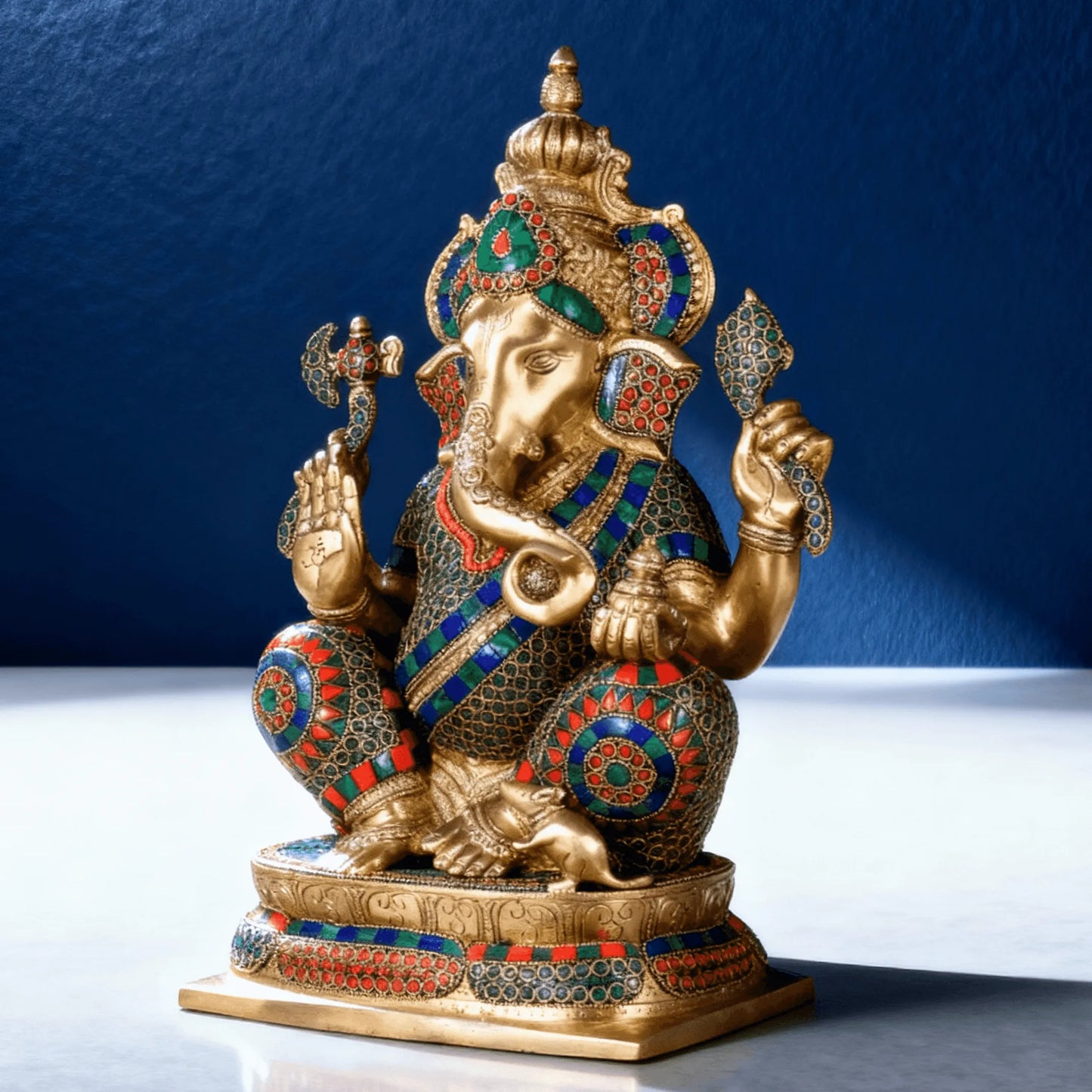 Handcrafted brass Lord Ganesha idol with green red and blue stone detailing, three-quarter rear angle on reflective surface