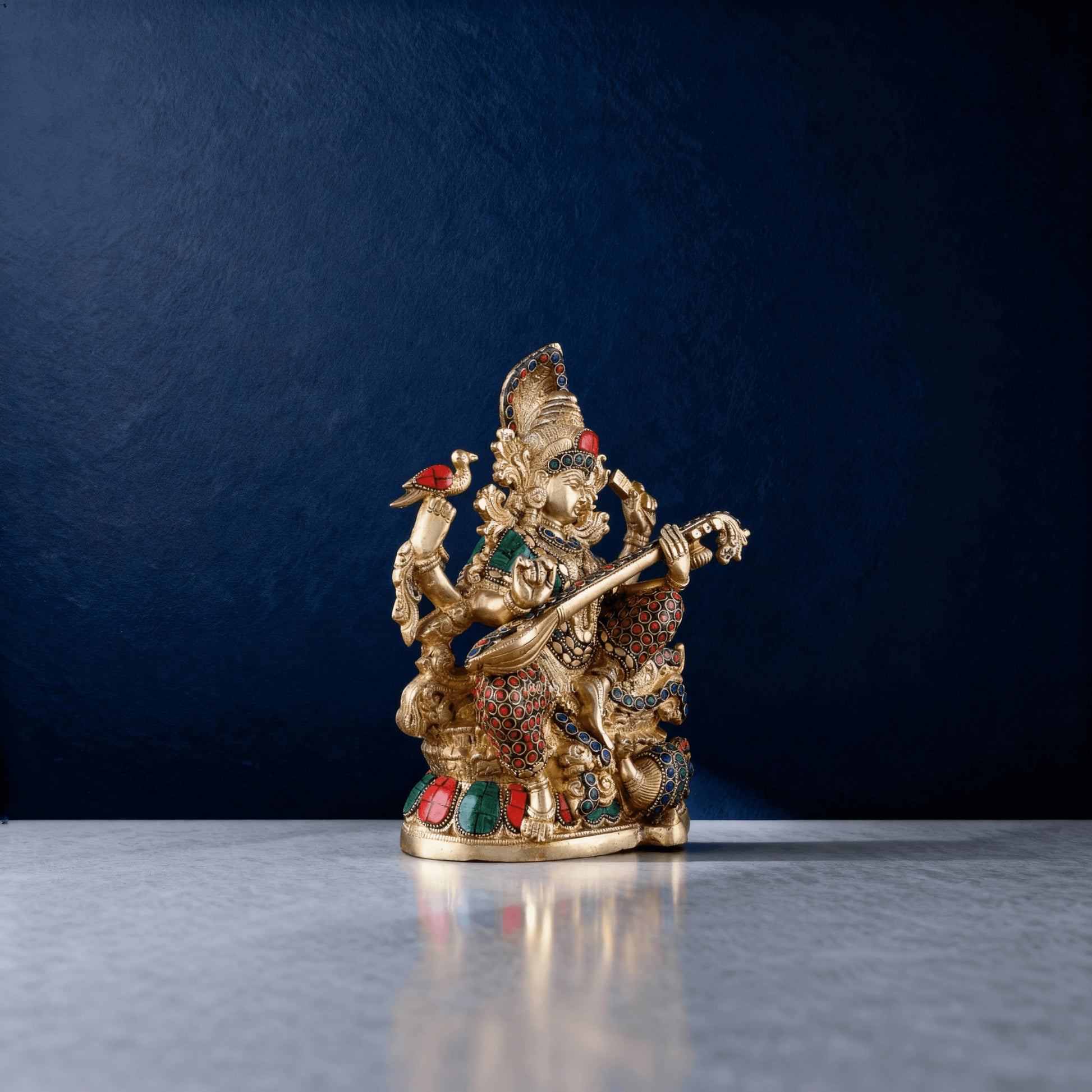 side view of brass Saraswati idol featuring detailed stonework and veena carving, placed on a reflective marble surface