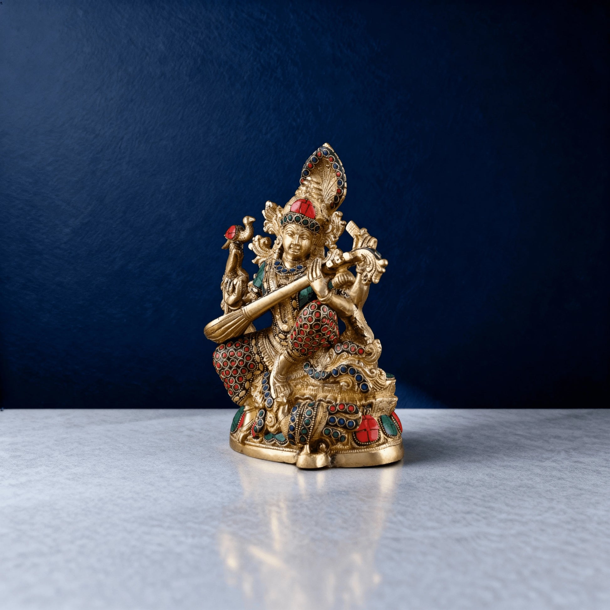 handcrafted Saraswati statue in brass with red and green stone inlay, shown in a front-angled view against a deep blue backdrop
