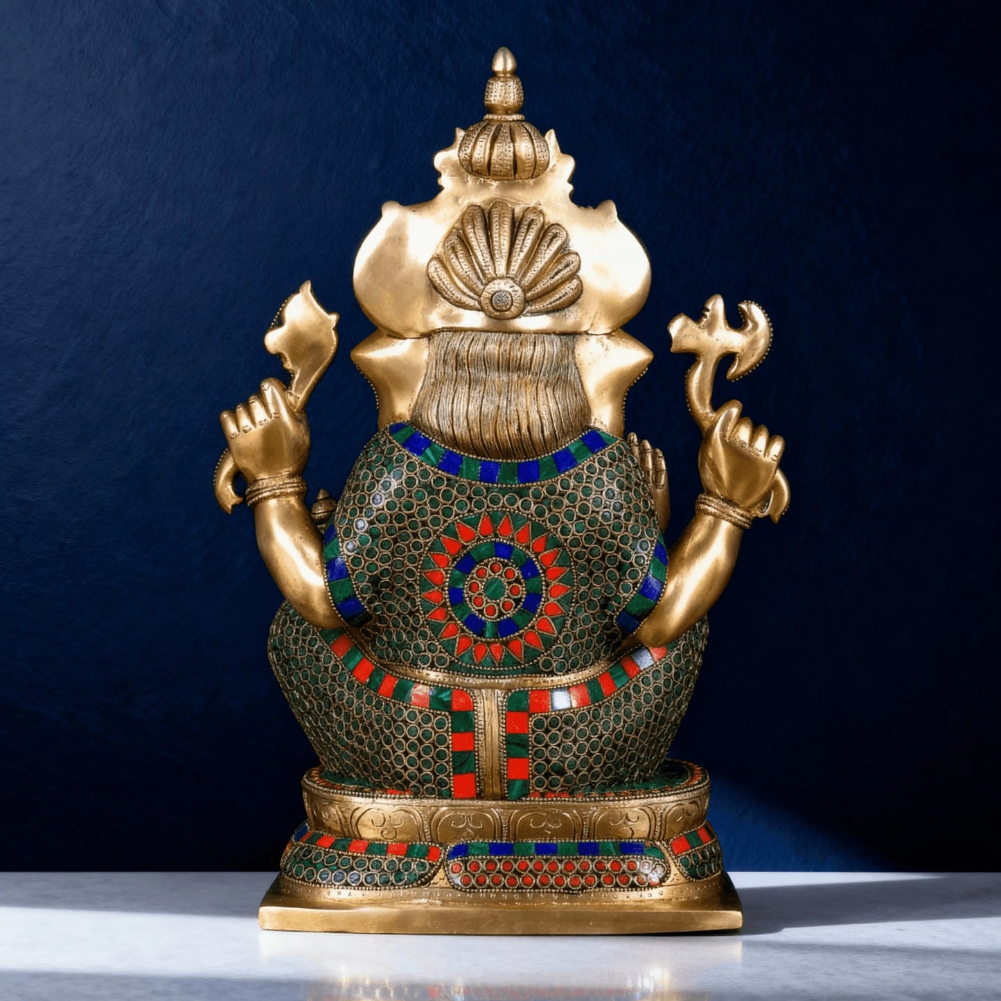 Brass Ganesha statue with colorful stone inlay, rear view showing intricate mosaic work and ornate crown against deep blue background