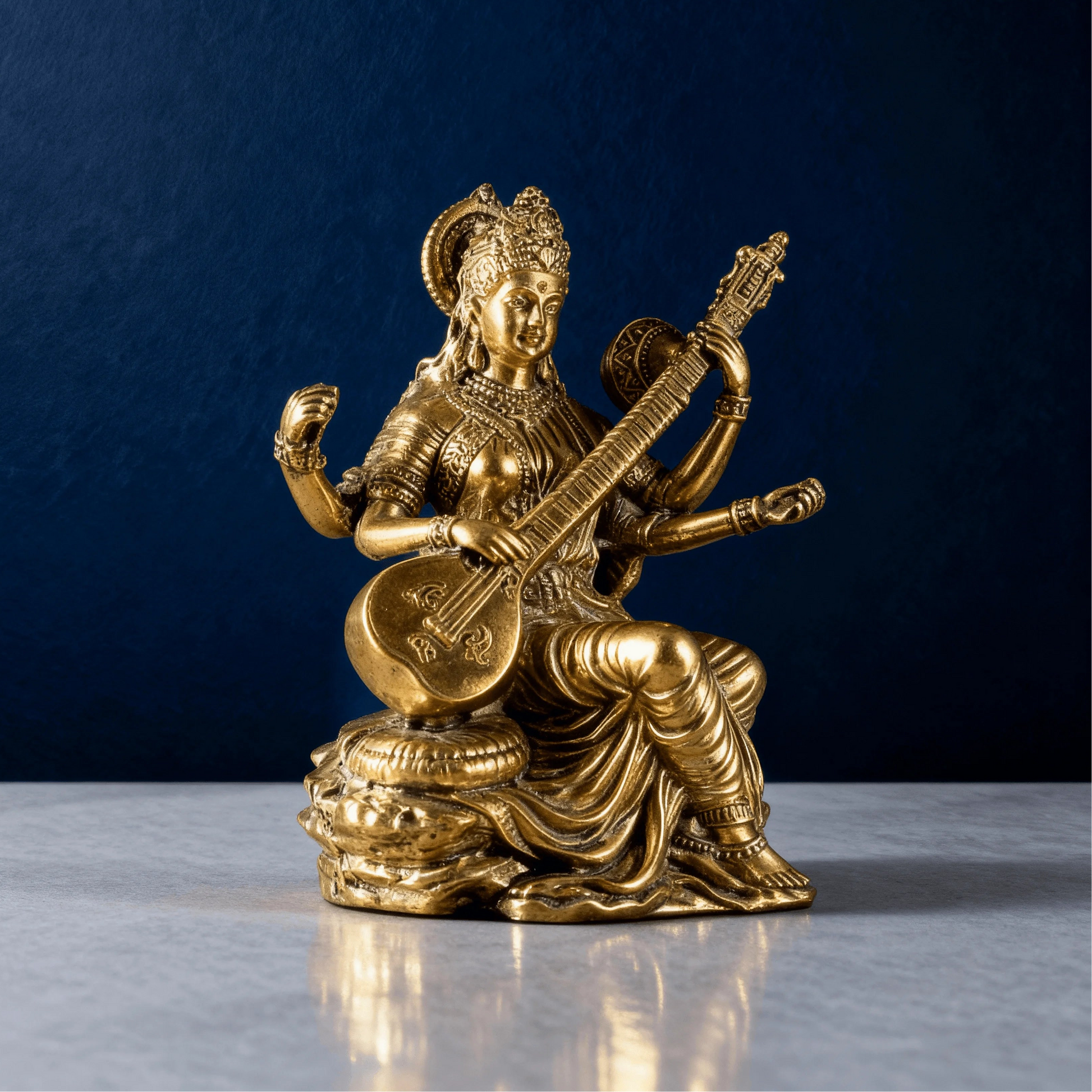 angled side view of brass Saraswati statue holding a veena, placed on a marble surface with a dark blue studio background